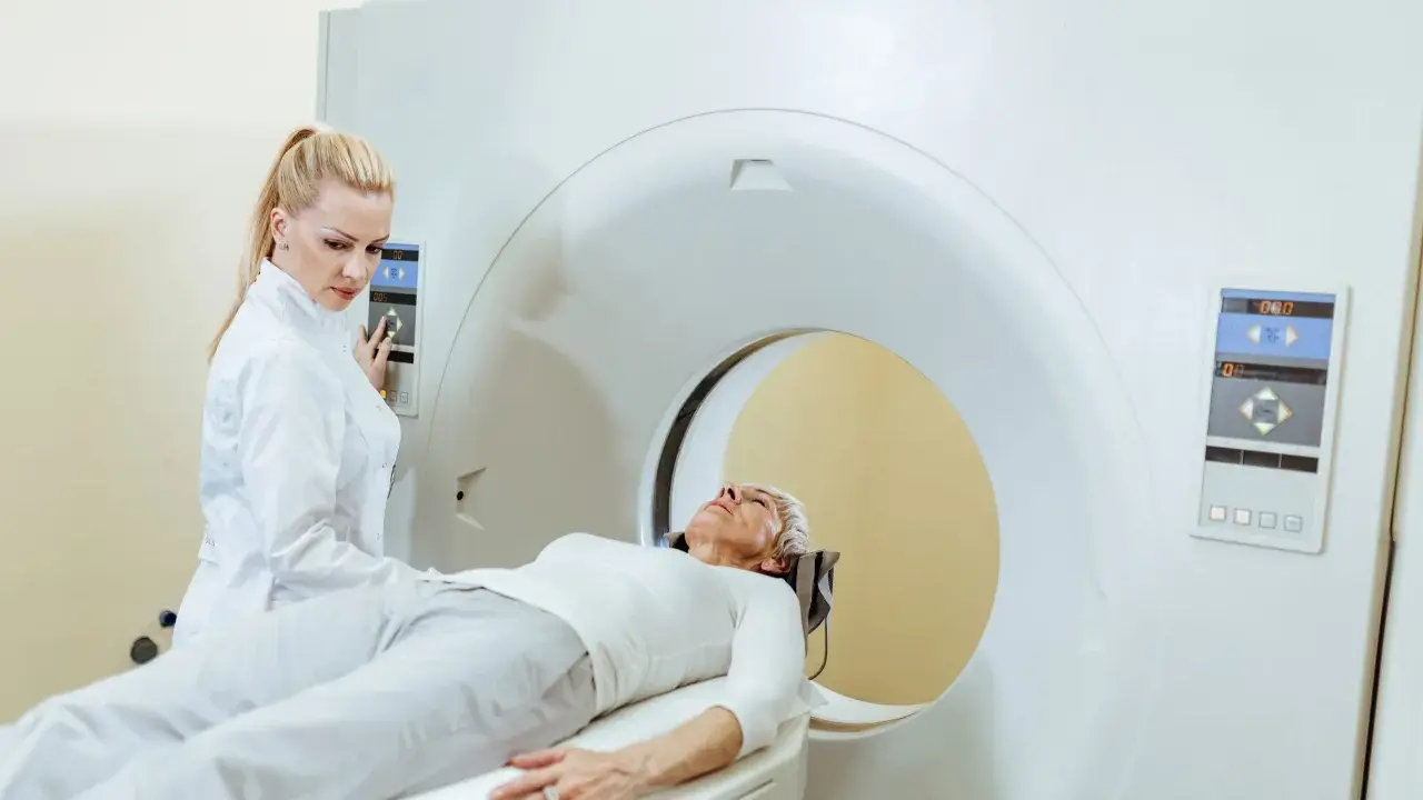 Kobieta w białej bluzie leży na łóżku rezonansu magnetycznego. Technik w białym fartuchu obsługuje panel sterowania.
