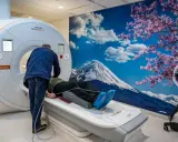 Pacjent leży na łóżku rezonansu magnetycznego, przygotowywany do badania. Za nim ściana z widokiem na górę Fudżi i kwitnącą wiśnię, tworząc silne wzmocnienie kontrastowe.