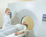 Kobieta w białej bluzie leży na łóżku rezonansu magnetycznego. Technik w białym fartuchu obsługuje panel sterowania.