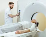 Pacjentka leży na łóżku aparatu do rezonansu magnetycznego, a technik przygotowuje badanie. Czy do rezonansu magnetycznego trzeba się rozebrać? Zazwyczaj tak, aby zdjąć metalowe przedmioty.
