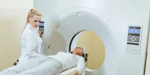 Kobieta w białej bluzie leży na łóżku rezonansu magnetycznego. Technik w białym fartuchu obsługuje panel sterowania.