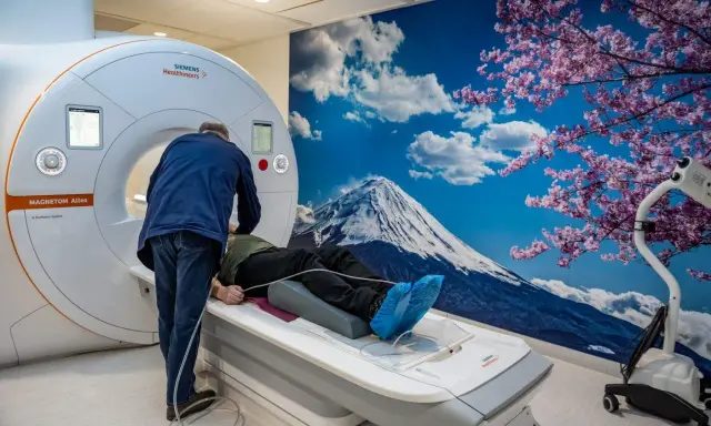 Pacjent leży na łóżku rezonansu magnetycznego, przygotowywany do badania. Za nim ściana z widokiem na górę Fudżi i kwitnącą wiśnię, tworząc silne wzmocnienie kontrastowe.
