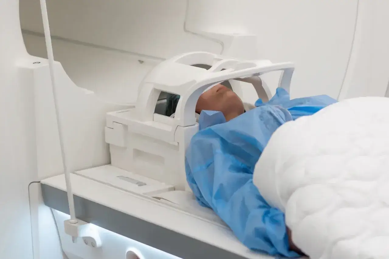 Pacjent w niebieskim fartuchu leży w aparacie do rezonansu magnetycznego głowy. Opis rezonansu magnetycznego głowy.