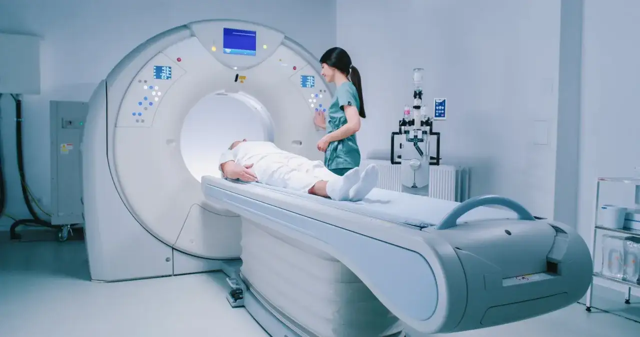 Pacjent leży na stole do rezonansu magnetycznego, ubrany w biały fartuch. Pielęgniarka przygotowuje badanie. Czy do rezonansu magnetycznego trzeba się rozebrać? Zazwyczaj tak, aby nic nie zakłócało obrazowania.