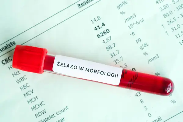 Probówka z krwią na tle wyników morfologii. Etykieta "ŻELAZO W MORFOLOGII" sugeruje badanie pod kątem przyczyn niedoboru żelaza.